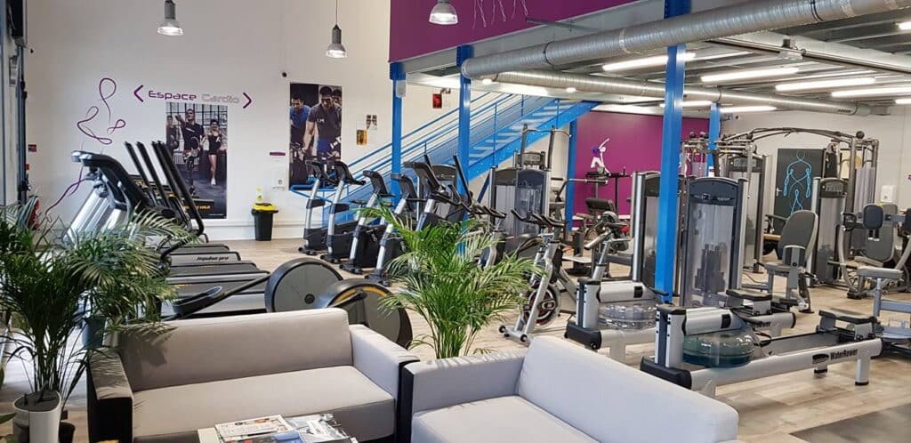 Salle de sport moderne avec machines cardio (tapis Impulse pro, rameurs WaterRower), espace musculation et coin détente.