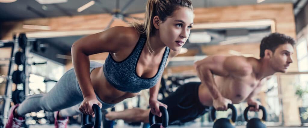 Femme et homme concentrés, faisant des pompes sur des kettlebells dans une salle de sport. Elle porte un débardeur de sport gris, lui est torse nu. Entraînement fitness.