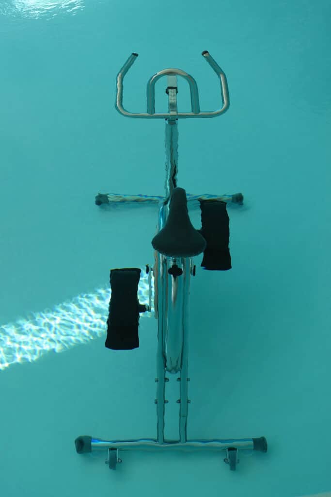 Aquabike sous l'eau dans une piscine turquoise, reflets lumineux sur le cadre métallique et les pédales noires.