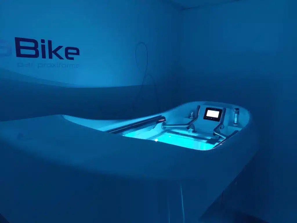 Machine d'hydro-exercice aquatique blanche illuminée en bleu, avec écran et poignées, sous l'inscription 'Bike par proxiforme' sur un mur bleu.