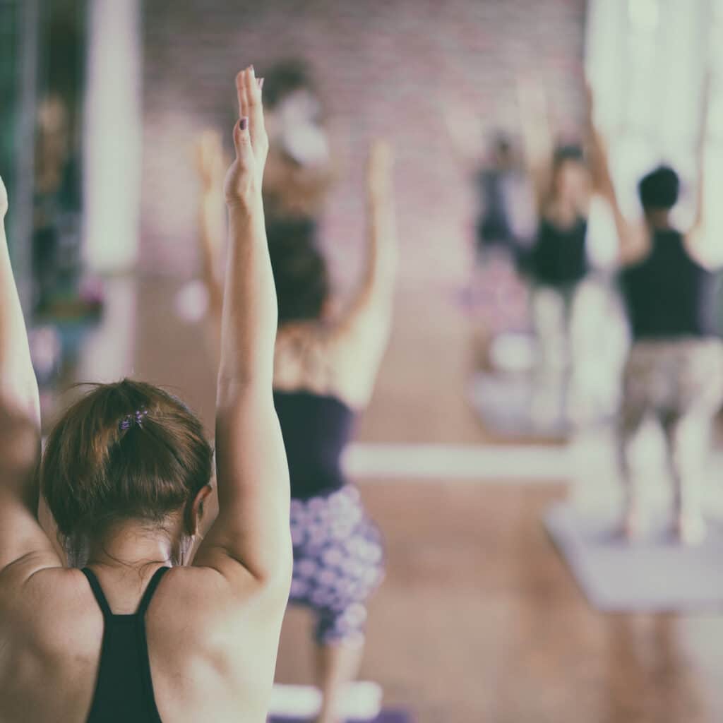 Femme en cours de yoga, vue de dos, bras levés en pleine pose. Autres yogis flous pratiquent en arrière-plan.