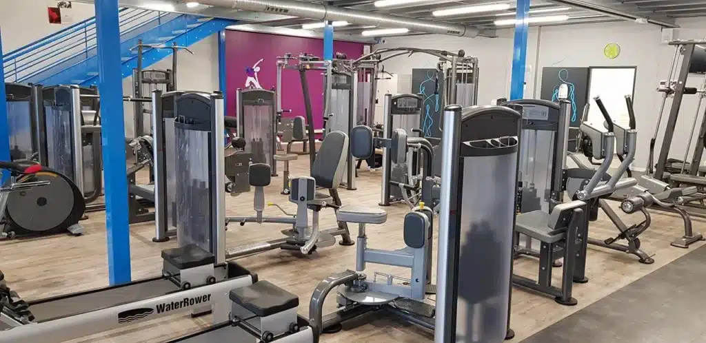 Salle de sport moderne avec escalier bleu, machines de musculation variées, rameur WaterRower et équipement cardio. Murs violet et gris décorés.